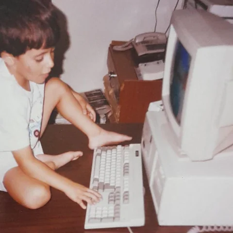 Mahdi at around 8, sitting on a desk and typing on a beige desktop computer.