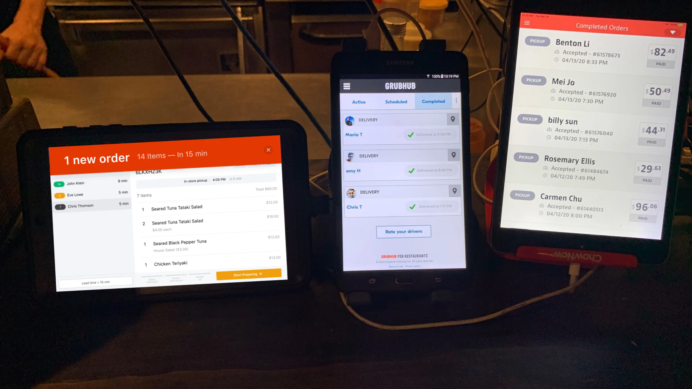 Three order tablets side by side on a restaurant counter at night: our console with a new team order, Grubhub's completed deliveries and ChowNow's completed pickups.