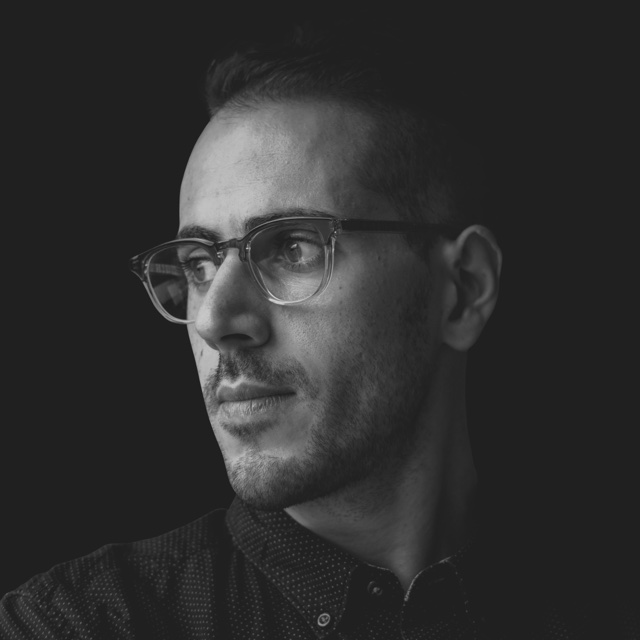 Black and white portrait of Mahdi Farra in glasses, looking to the side, on a dark background.