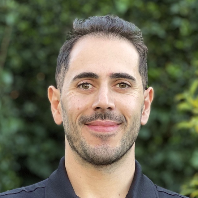 Mahdi Farra smiling outdoors in front of green leaves, wearing a dark shirt.